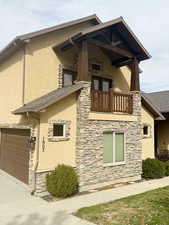 View of side of property with a balcony, stucco siding, stone siding, driveway, and an attached garage