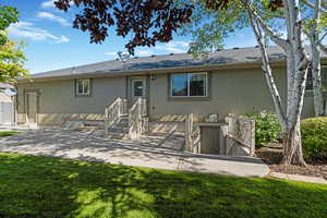 Back of house with shingled roof, view of patio and barbeque area, wide stairway leading to basement entrance, lawn, and stucco siding