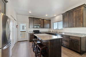 Kitchen featuring dark brown cabinets, stainless steel appliances, light stone counters, light wood-style flooring, and a kitchen island