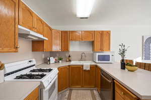Kitchen with white appliances, brown cabinetry, under cabinet range hood, light countertops, and a peninsula
