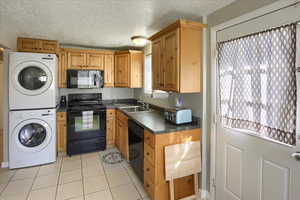 Kitchen featuring black appliances, dark countertops, light tile patterned floors, a textured ceiling, and stacked washer / dryer