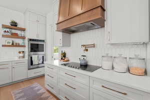 Kitchen featuring decorative backsplash, light stone countertops, double oven, custom exhaust hood, and white cabinets