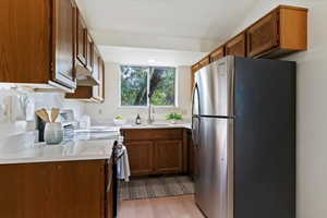 Kitchen with appliances with stainless steel finishes, brown cabinetry, light wood finished floors, under cabinet range hood, and light stone countertops