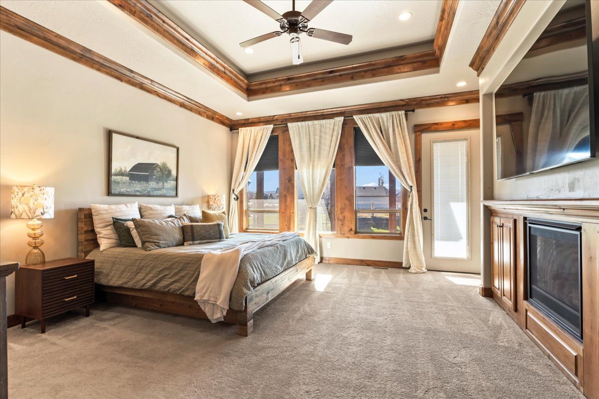 Primary Bedroom with light carpet, a raised ceiling, a glass covered fireplace, ornamental molding, and ceiling fan