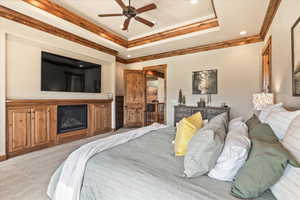 Carpeted bedroom with a raised ceiling, crown molding, ceiling fan, and a glass covered fireplace