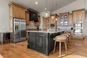 Kitchen with two tone color scheme, wine cooler, stainless steel appliances, a kitchen island with sink, and light stone countertops