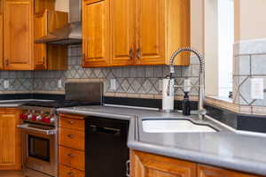 Kitchen with wall chimney range hood, brown cabinetry, premium range, backsplash, and black dishwasher
