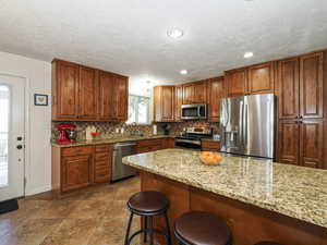 Kitchen with stainless steel appliances, decorative backsplash, a kitchen breakfast bar, brown cabinetry, and light stone counters