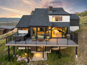 Back of property at dusk with a hot tub, a wooden deck, a shingled roof, a chimney, and board and batten siding