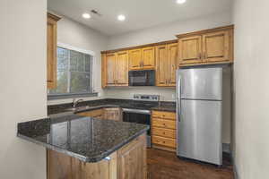 Kitchen with stainless steel appliances, a peninsula, brown cabinetry, dark wood-style floors, and dark stone counters