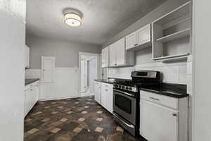 Kitchen featuring dark countertops, stainless steel range with gas stovetop, white cabinetry, a textured ceiling, and a wainscoted wall