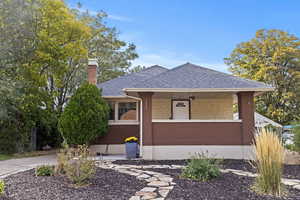 View of front of home featuring a shingled roof, a chimney, covered porch, and brick siding