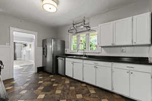 Kitchen with white cabinets, a textured ceiling, appliances with stainless steel finishes, dark stone finish floors, and a wainscoted wall