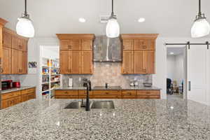 Kitchen featuring brown cabinets, a barn door, decorative light fixtures, backsplash, and wall chimney range hood