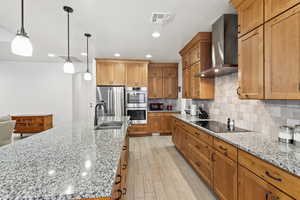Kitchen featuring appliances with stainless steel finishes, wall chimney range hood, light stone countertops, decorative light fixtures, and brown cabinetry