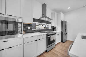 Kitchen with white cabinets, appliances with stainless steel finishes, wall chimney range hood, tasteful backsplash, and light wood finished floors