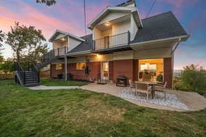 Back of property at dusk with stairway, a patio, a lawn, a shingled roof, and brick siding