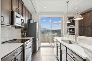 Kitchen with appliances with stainless steel finishes, beamed ceiling, dark brown cabinetry, light stone countertops, and light wood-style flooring