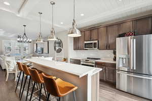 Kitchen featuring beamed ceiling, appliances with stainless steel finishes, tasteful backsplash, pendant lighting, and dark brown cabinets