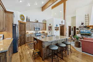 Kitchen with beamed ceiling, light stone counters, light wood-style floors, a center island with sink, and open floor plan