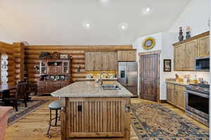 Kitchen with vaulted ceiling, appliances with stainless steel finishes, light wood-style floors, light stone counters, and a kitchen island with sink