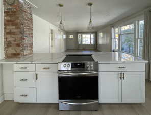 Kitchen with stainless steel electric range oven, white cabinets, light stone countertops, hanging light fixtures, and light wood-style flooring
