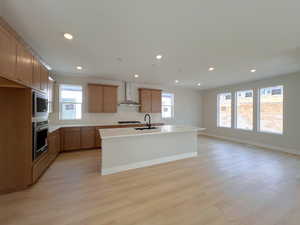 Kitchen featuring brown cabinets, recessed lighting, a center island with sink, stainless steel appliances, and wall chimney range hood