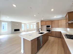 Kitchen featuring light wood-type flooring, a tile fireplace, an island with sink, stainless steel appliances, and brown cabinetry