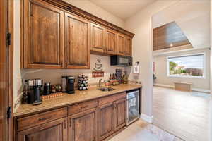 Kitchenet with wine cooler, light stone countertops, black microwave, brown cabinets, and light wood-style flooring