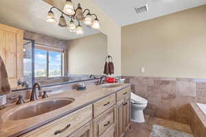 Bathroom featuring tile walls, a textured ceiling, double vanity, tiled bath, and wainscoting