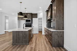 Kitchen with dark brown cabinets, hanging light fixtures, a kitchen island with sink, appliances with stainless steel finishes, and light stone counters