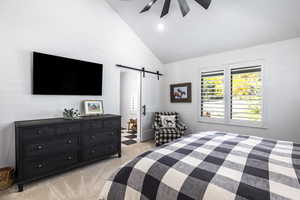Bedroom with a barn door, light colored carpet, high vaulted ceiling, connected bathroom, and a ceiling fan