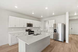 Kitchen featuring appliances with stainless steel finishes, white cabinetry, an island with sink, light stone countertops, and light wood-type flooring