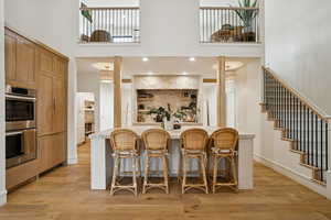 Kitchen featuring a breakfast bar area, a towering ceiling, stainless steel double oven, brown cabinets, and light wood-style flooring