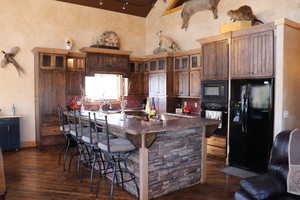 Kitchen with black appliances, a breakfast bar area, dark wood-style floors, a large island with sink, and glass insert cabinets