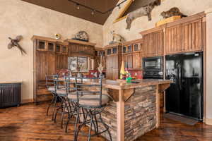 Kitchen with a kitchen breakfast bar, black appliances, glass insert cabinets, dark wood-style flooring, and high vaulted ceiling
