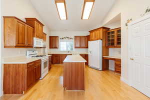 Kitchen featuring brown cabinets, white appliances, light countertops, vaulted ceiling, and glass insert cabinets