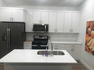 Kitchen featuring appliances with stainless steel finishes, dark wood-style flooring, white cabinetry, a center island with sink, and light stone countertops