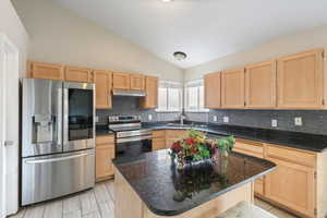 Kitchen featuring light brown cabinetry, appliances with stainless steel finishes, lofted ceiling, backsplash, and a kitchen island