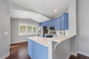 Kitchen with blue cabinets, vaulted ceiling, stainless steel appliances, backsplash, breakfast counter, and dark wood-style floors