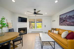 Living room with a fireplace, a ceiling fan, light LVP  flooring, recessed lighting, and a baseboard heating unit
