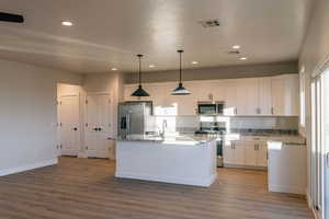 Kitchen with decorative light fixtures, appliances with stainless steel finishes, white cabinets, a center island with sink, and light stone counters