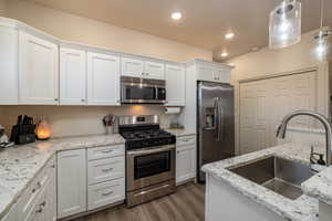 Kitchen with stainless steel appliances, white cabinetry, light wood-type flooring, light stone counters, and pendant lighting