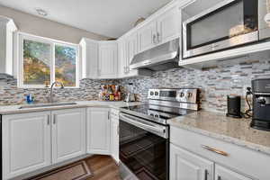 Kitchen with stainless steel appliances, white cabinets, light stone countertops, under cabinet range hood, and backsplash