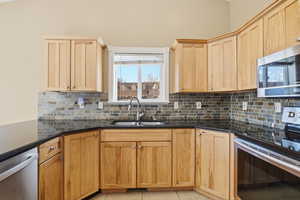 Kitchen with appliances with stainless steel finishes, backsplash, light brown cabinets, light tile patterned flooring, and dark stone counters