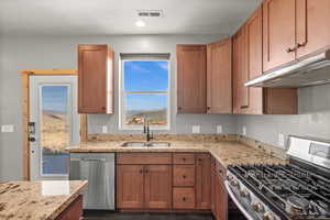 Kitchen featuring appliances with stainless steel finishes, light stone counters, brown cabinetry, under cabinet range hood, and a textured ceiling