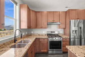 Kitchen with stainless steel appliances, light stone countertops, brown cabinetry, under cabinet range hood, and a textured ceiling