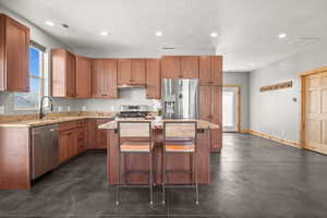 Kitchen featuring light stone counters, appliances with stainless steel finishes, brown cabinetry, a kitchen island, and recessed lighting