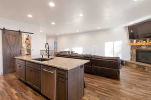 Kitchen featuring a barn door, dark brown cabinetry, light stone countertops, stainless steel dishwasher, and dark wood finished floors