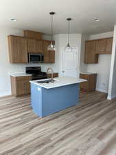 Kitchen featuring brown cabinets, pendant lighting, stainless steel appliances, an island with sink, and light wood finished floors
